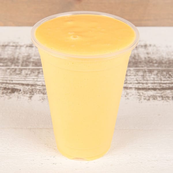 A vibrant yellow mango smoothie in a clear plastic cup on a light wooden surface.